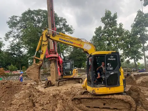 Ekskavator kuning dengan operator di kabin, di sebelahnya alat bor bertiang tinggi, di tanah galian