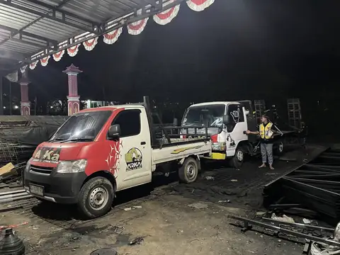 Dua pickup bergambar ARKA di dalam bangunan beratap seng pada malam hari, seorang pria berompi kuning berdiri di dekatnya