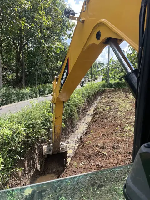 Lengan ekskavator kuning menggali parit sempit di tepi jalan, dilihat dari kabin