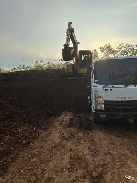 Ekskavator di atas tanah gelap yang sudah digali, dengan truk Isuzu putih di sampingnya saat matahari terbenam