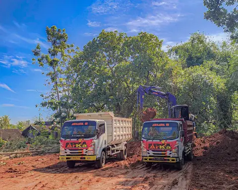 Dua dump truk bergambar ARKA di tanah merah, dengan ekskavator biru dan pepohonan di belakangnya