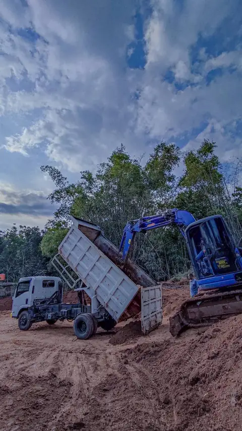 Dump truk menaikkan bak untuk menurunkan tanah di samping ekskavator biru