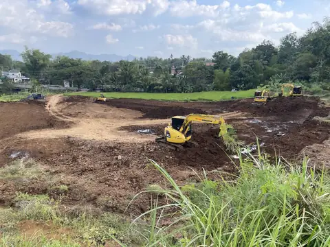 Ekskavator kuning di lahan galian yang luas, dengan alat lain di kejauhan dan perbukitan di belakang