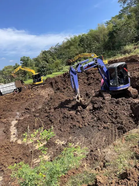 Ekskavator biru menggali tanah di lereng, dengan ekskavator kuning di belakangnya