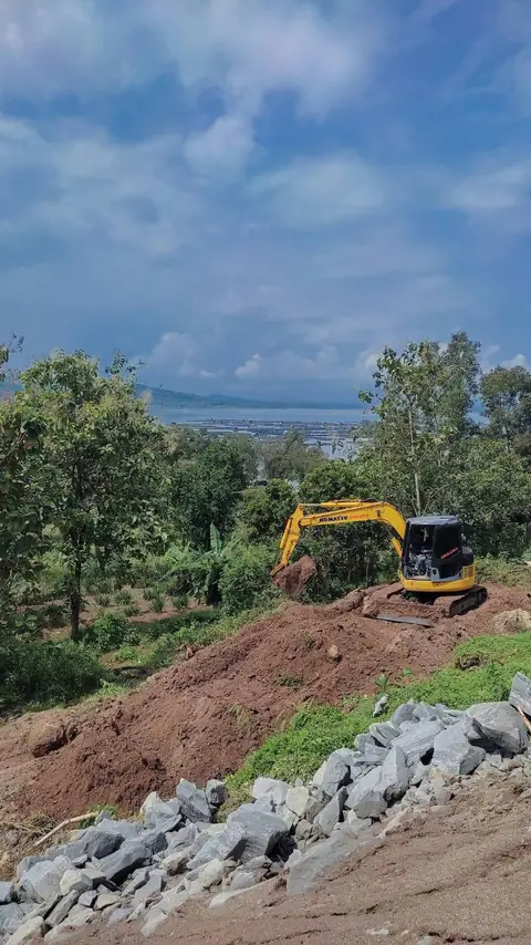 Ekskavator kuning di atas gundukan tanah di lereng, dengan tumpukan batu besar di bagian depan