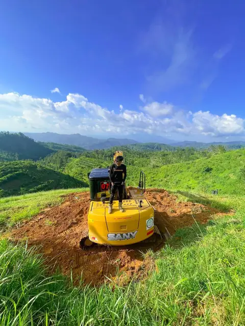 Seorang pria berdiri di atas ekskavator kuning di tanah terbuka di puncak bukit berumput