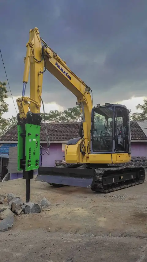 Ekskavator kuning dengan breaker hijau di lengannya, di samping rumah-rumah