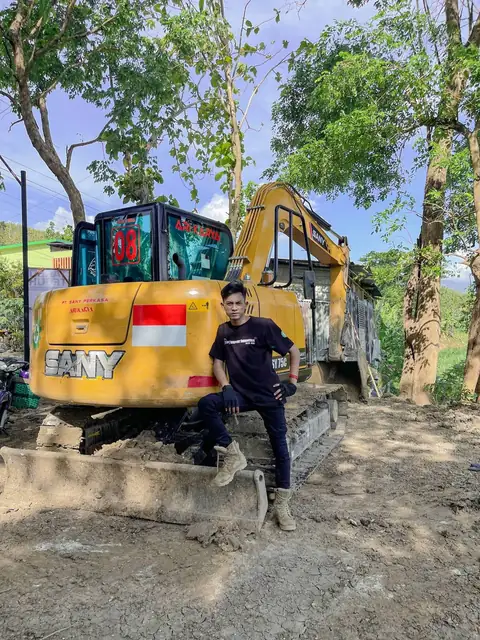 Seorang pria berdiri di samping ekskavator kuning bertulisan SANY, dengan bendera kecil di kabinnya