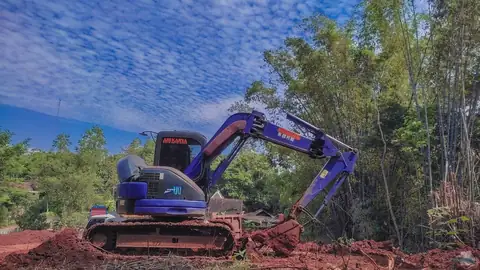 Ekskavator biru kecil di tanah merah, dengan pepohonan dan langit sore di belakangnya
