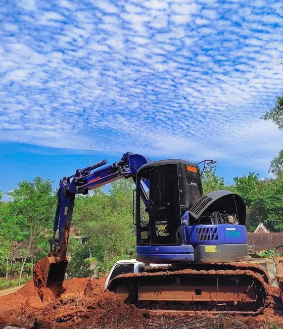 Ekskavator biru di tanah merah dengan langit berawan