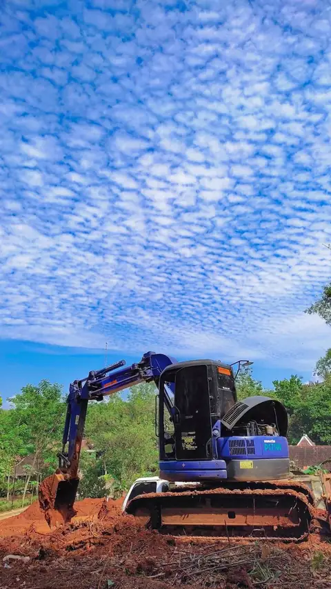 Ekskavator biru di tanah merah dengan langit penuh awan tipis
