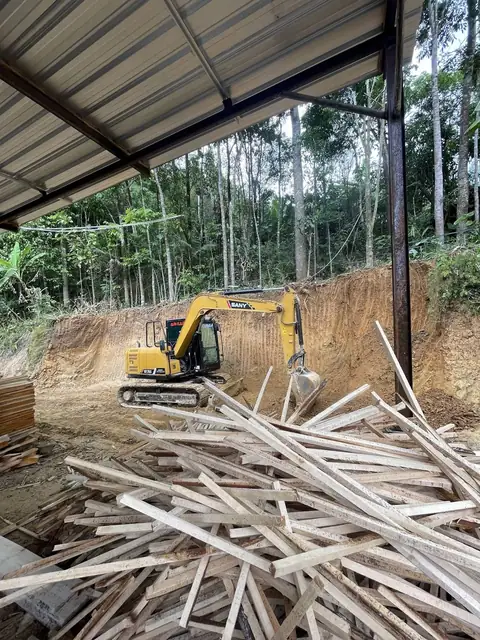 Ekskavator kuning menggali tebing tanah di bawah atap seng, dengan tumpukan papan kayu di depannya