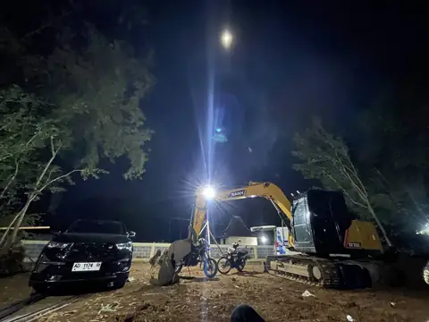 Ekskavator kuning bekerja malam hari dengan lampu sorot terang, sebuah mobil hitam terparkir di dekatnya