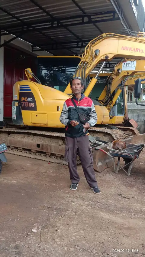 Seorang pria berdiri di depan ekskavator kuning di dalam bangunan beratap seng
