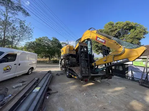 Ekskavator kuning di atas truk pengangkut, dengan besi panjang di tanah dan sebuah van putih di belakangnya