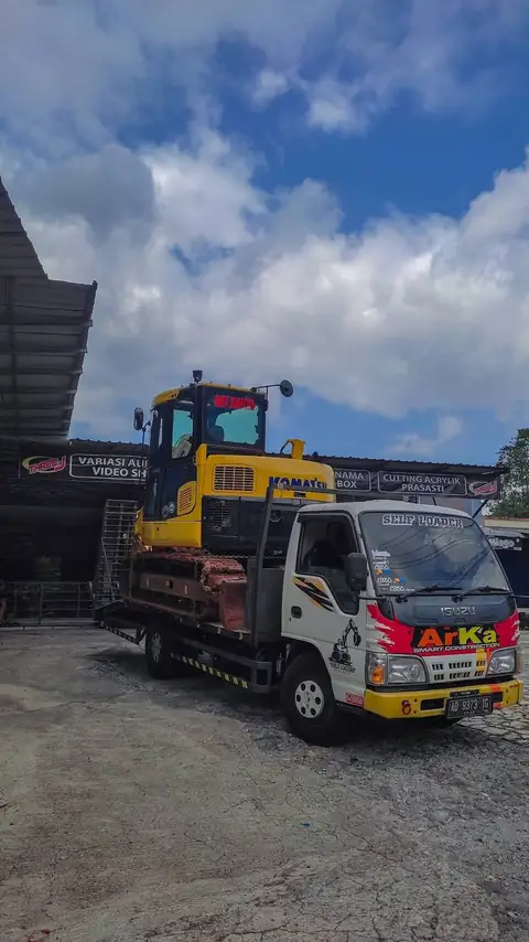 Ekskavator kuning di atas truk di halaman berbatu, langit biru berawan