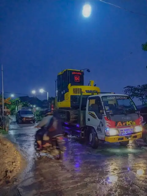 Truk ARKA membawa alat kuning di jalan basah saat senja, dengan lampu jalan menyala