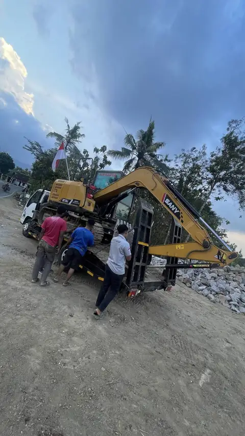 Tiga orang berdiri di dekat ekskavator kuning yang berada di atas truk pengangkut