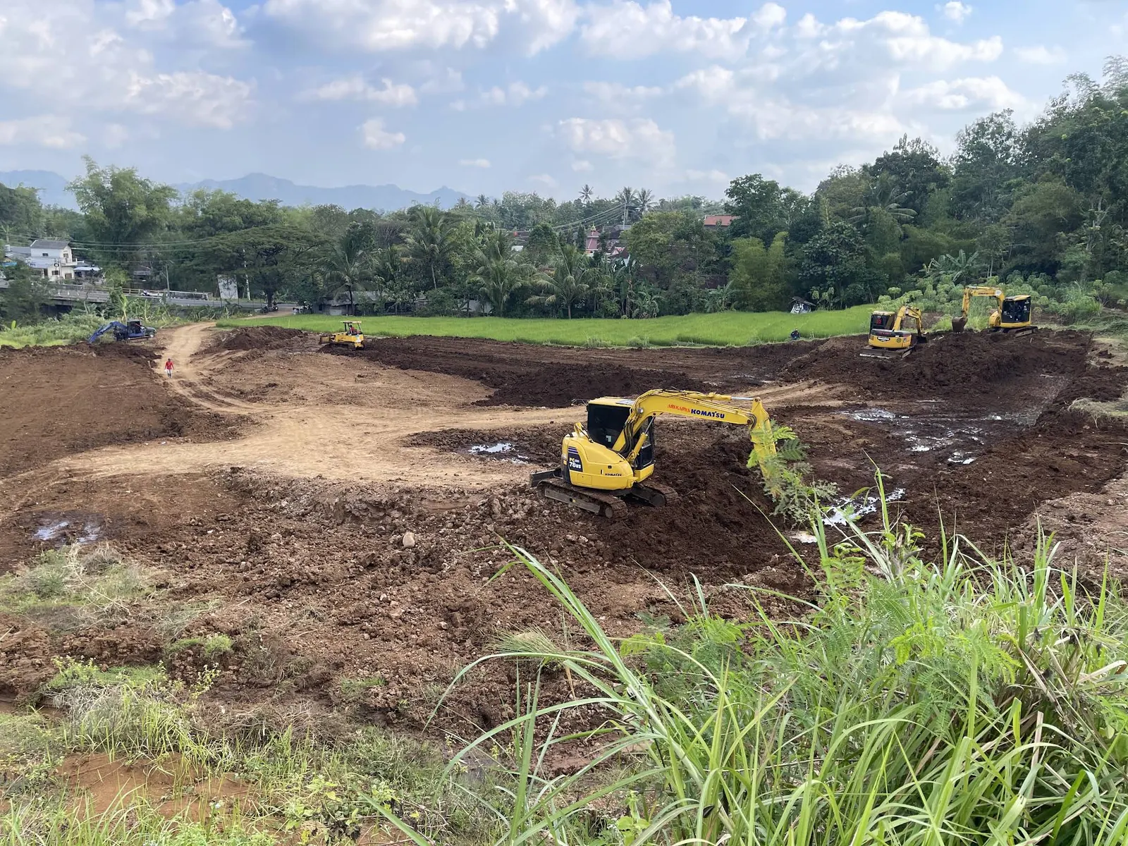 Ekskavator kuning di lahan galian yang luas dengan perbukitan di belakangnya